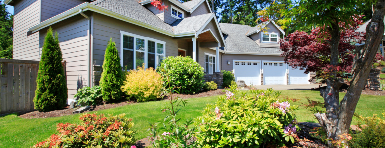 Large beige sided house with white trim and shutters and a large green grass front yard with a lot of colorful flowers and trees