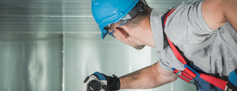 Caucasian male with blue hard hat and maintenance gloves on using a flashlight to look into an air duct