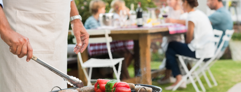 Man standing in front of an outdoor grill holding utensils barbecuing food in the backyard with people sitting at a table next to him