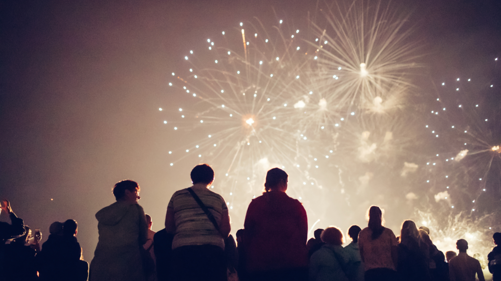 People sitting outside in the dark looking up at a fireworks display in the sky