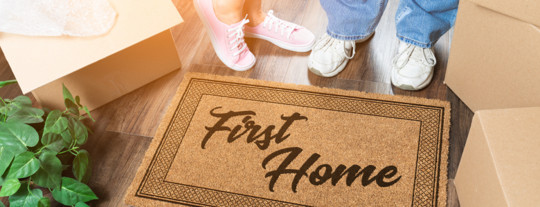 two people standing in fron of a door mat that reads first home with a green plant to their left and showing only their feet