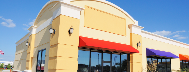 Beige commercial building for sale with red awning and bright blue sky glass windows and door with black trim to highlight commercial real estate