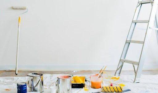 White wall with a paint roller leaning against it, ladder up, floor covering and paint supplies sitting on the floor