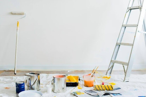 White wall with a paint roller leaning against it, ladder up, floor covering and paint supplies sitting on the floor