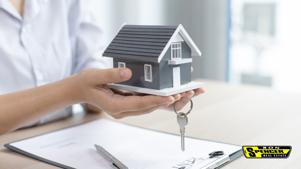 Caucasian person holding a model of a gray home and a house key at a desk with mortgage paperwork