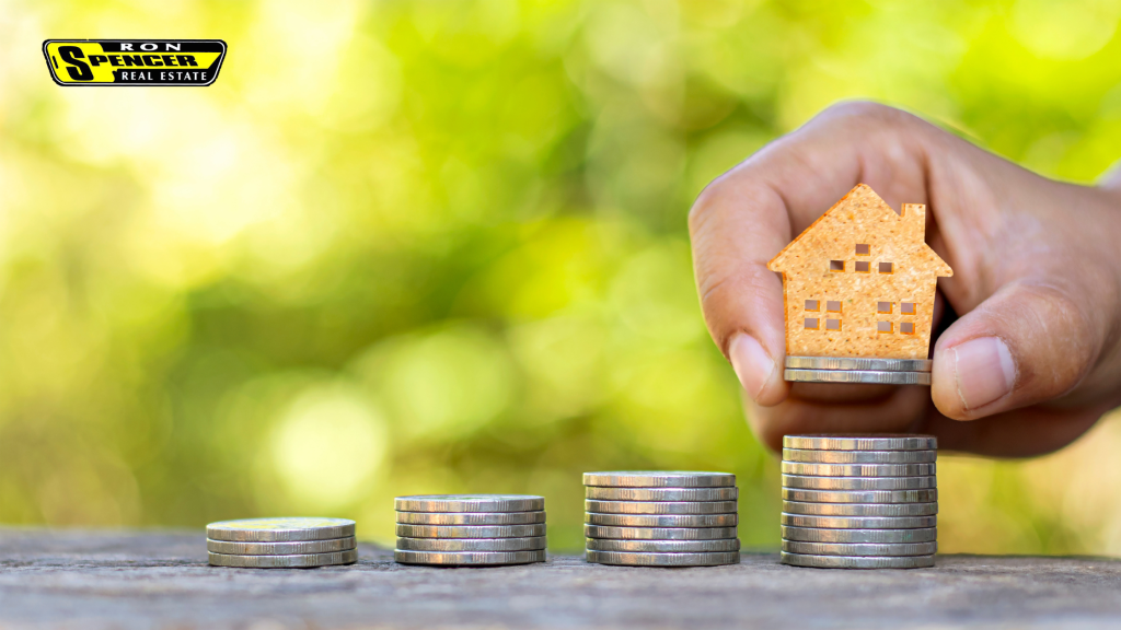 Outdoor green foliage background with a hand holding a mini home model sitting it on top of various stacks of coins