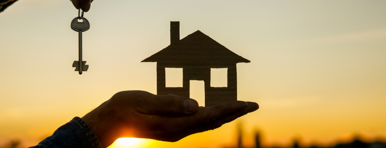 A hand holding a key and house cut out in front of a sunset and outside scenery