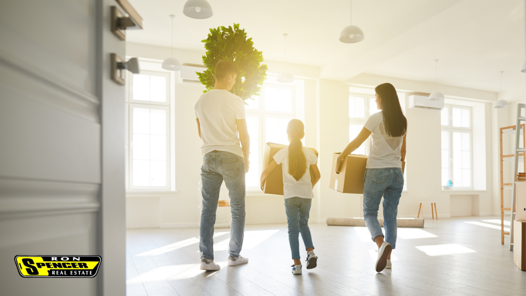 Man woman and child carrying moving boxes and a green plant to the door with sun shining in the windows