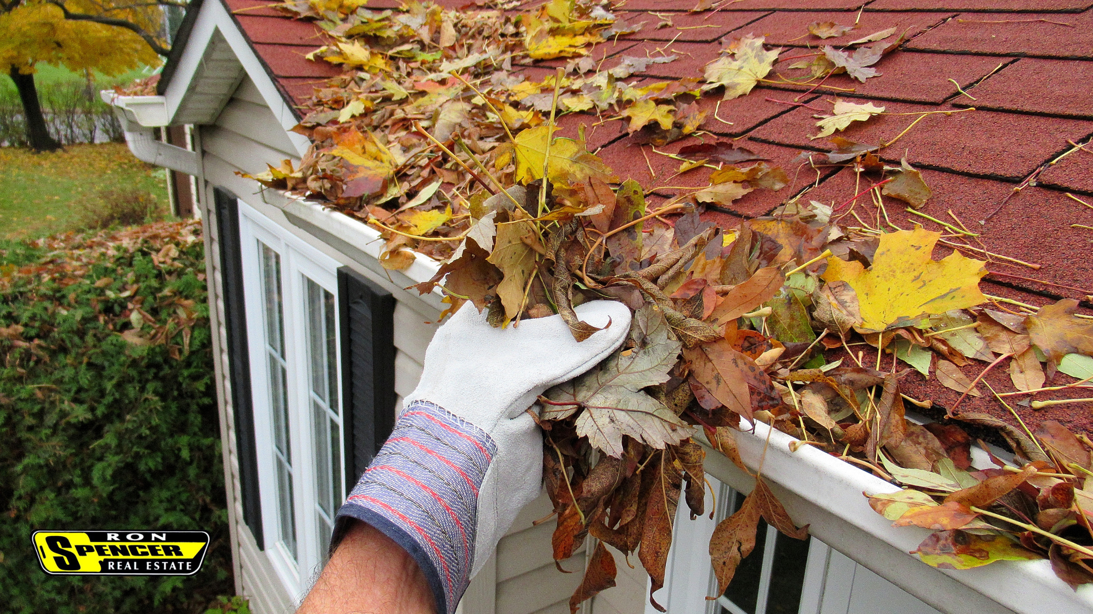 A gloved hand cleaning autumn colored leaves in gold and red and orange off of a roof with red shingles and gray siding