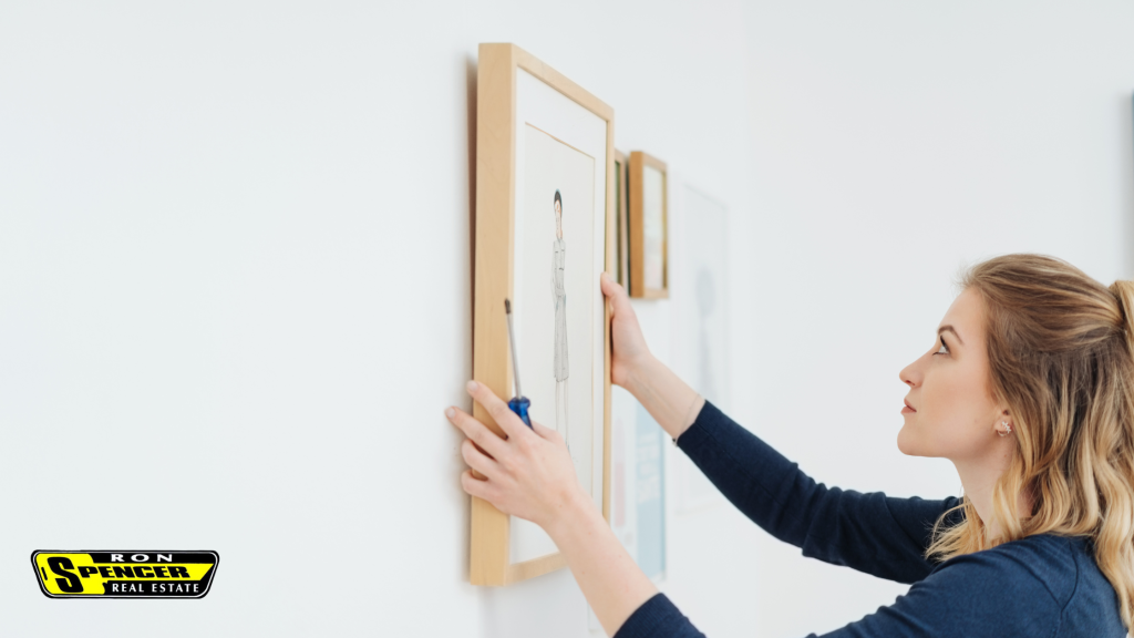 a caucasian woman with blonde hair in a black top hanging a piece of artwork in a blonde wood frame on a white wall lining it up to hang properly
