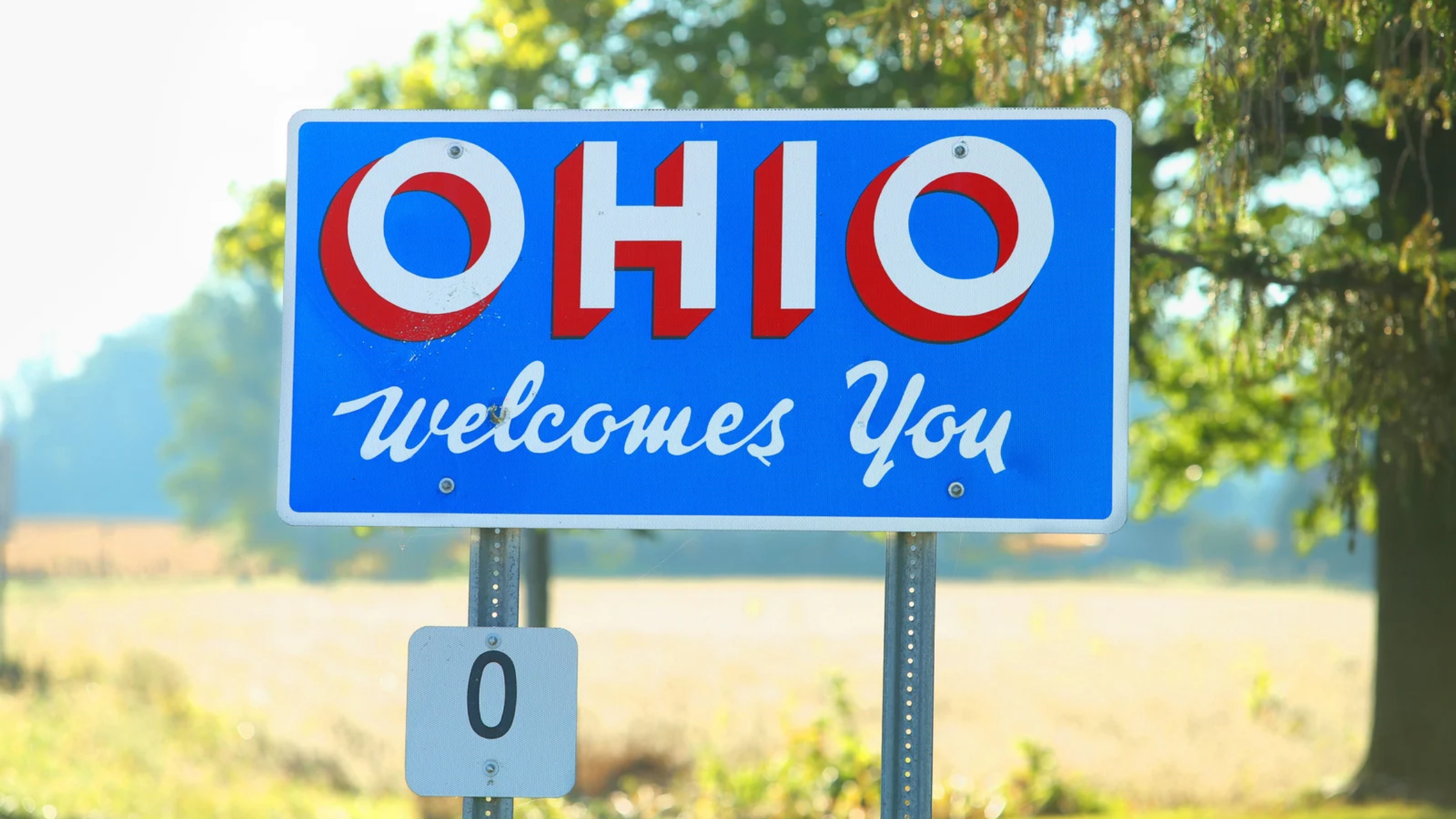 a rural scene with a highway sign that reads: Ohio Welcomes You in red and white letters on a blue background